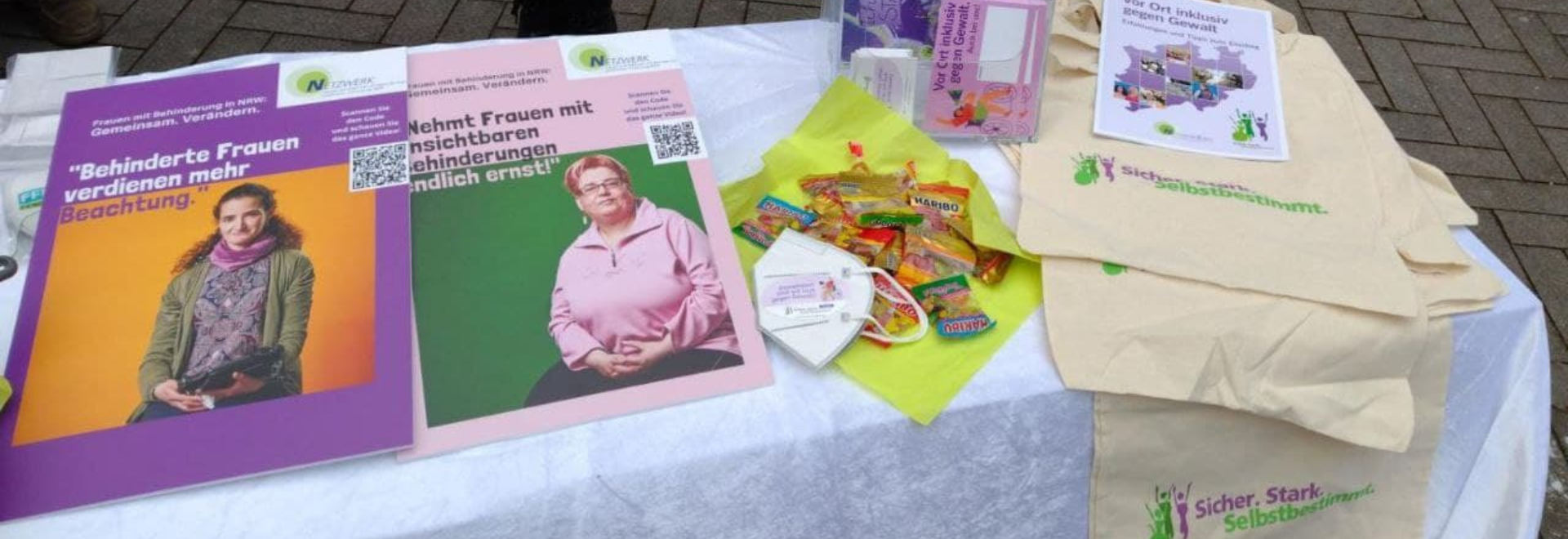 Auf dem Bild sieht man einen Informationstisch mit verschiedenen Materialien. Es gibt zwei große Plakate, die sich auf die Rechte von Frauen mit Behinderungen konzentrieren und Netzwerk-Frauen zeigen. Auf dem Tisch liegen außerdem einige Werbeartikel wie Süßigkeiten, Flyer, FFP2-Masken und Stofftaschen mit dem Slogan "Sicher. Stark. Selbstbestimmt."