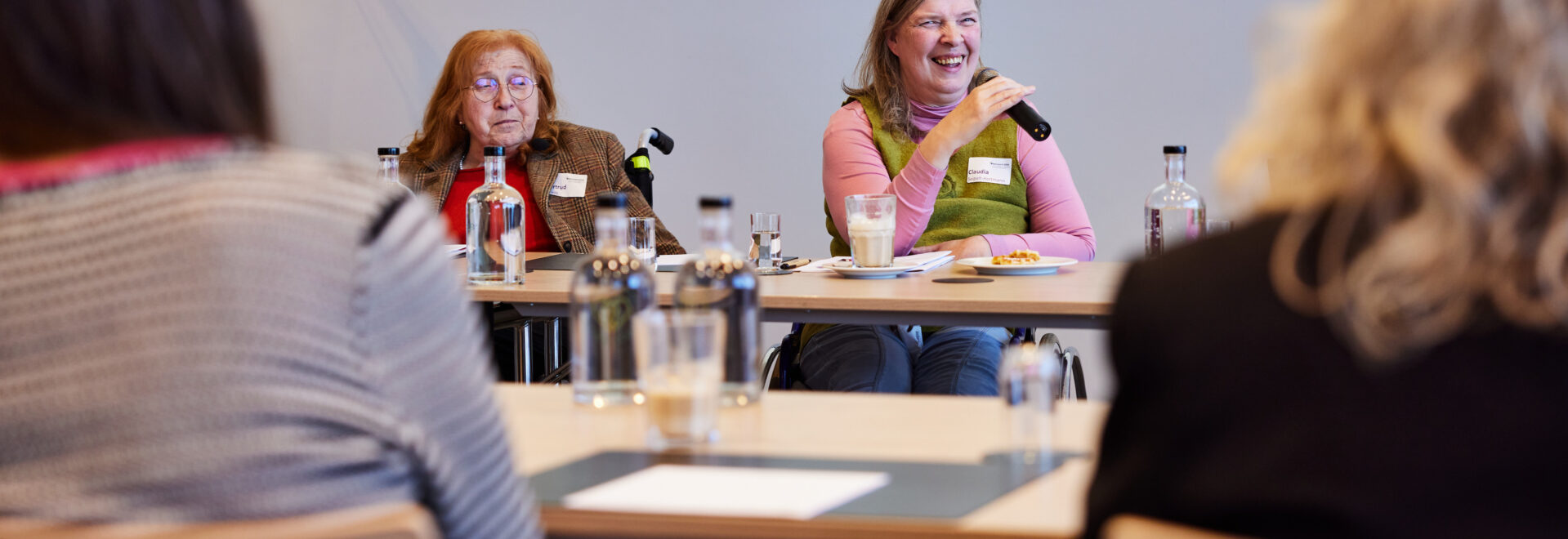 Bick durch Stuhlreihen auf zwei Frauen, die an einem Tisch vor Kopf sitzen, beide im Rollstuhl. Eine hat rote Haare und eine Brille. Die Frau rechts hat lange grau-blonde Haare und spricht in eine Mikrofon.