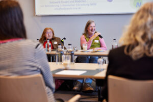 Bick durch Stuhlreihen auf zwei Frauen, die an einem Tisch vor Kopf sitzen, beide im Rollstuhl. Eine hat rote Haare und eine Brille. Die Frau rechts hat lange grau-blonde Haare und spricht in eine Mikrofon.