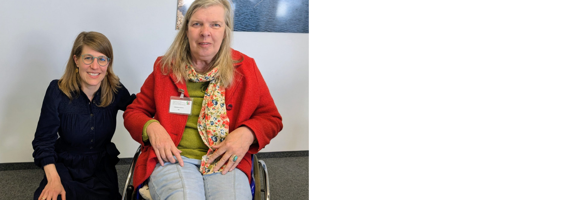 Zwei Frauen mit langen Haaren lächeln in die Kamera. Die linke Frau, im blauen Kleid, trägt eine Brille. Sie kniet. Die rechte Frau, im roten Blazer, sitzt im Rollstuhl.